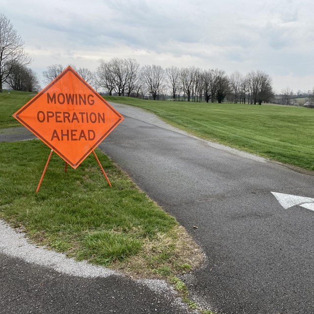 An orange caution sign labeled Warning Mowers Ahead is posted in a parkinglot.