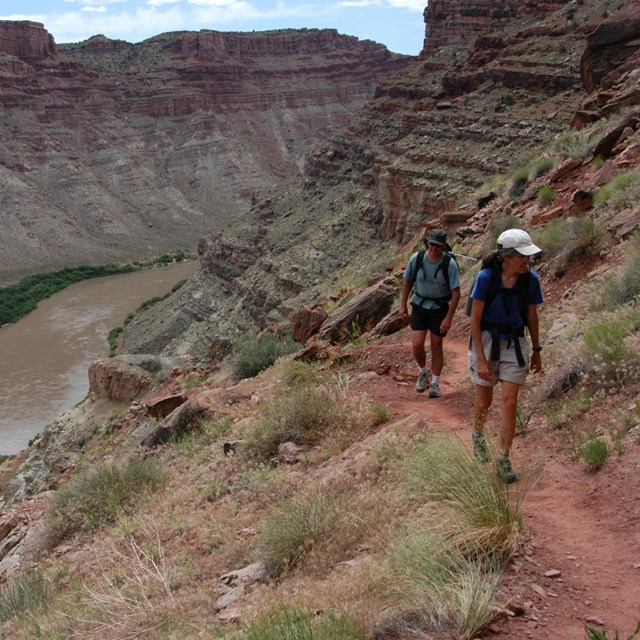 two hikers walk on a trail with a river below