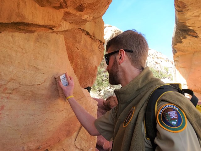 A person in a tan uniform and sunglasses brushing a sandstone wall.