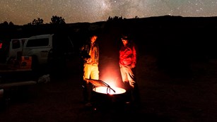 Two people stand beside a campfire, with the Milky Way above them. 