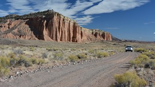 White SUV on gravel road, with red cliffs and blue skies and clouds in the background.
