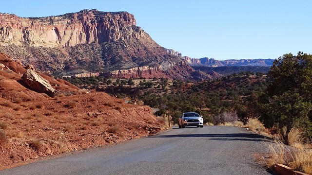 A car drives a narrow road with tall red cliffs rising on one side of the road.
