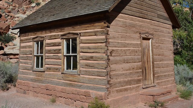 Small log cabin-like building with windows and a door, below red rock cliffs and blue sky.