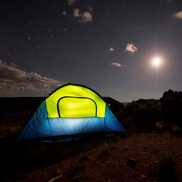 Green and blue tent with a light inside set up at night, with dark skies and a full moon above.