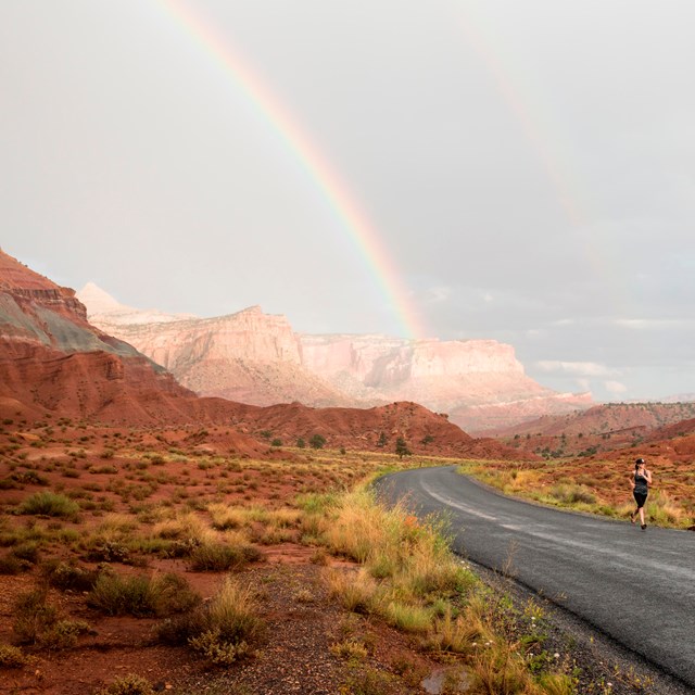 A woman runs on a paved road near towering red cliffs. A rainbow arcs across the gray sky.