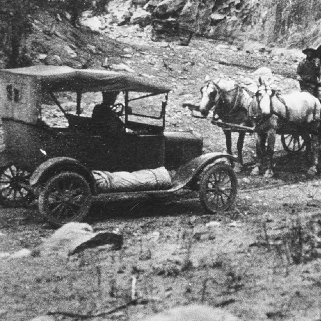 Black and white photo of old fashioned car meeting two wagons pulled by horses in a narrow canyon.