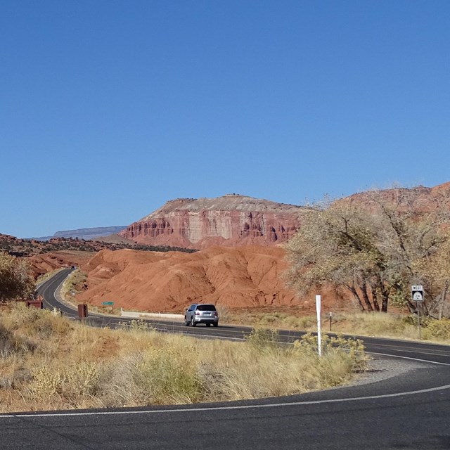 Stone building blends into red rocks around it, with blacktop road, car, some shrubs, and blue skies