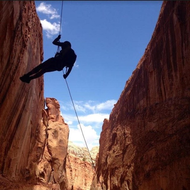 Silhouette of person on rope, between cliffs, with blue sky in the background.