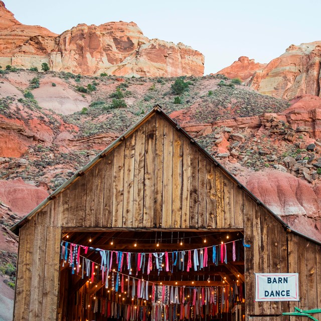 Large wooden barn below red cliffs, with festive decorations and people celebrating.