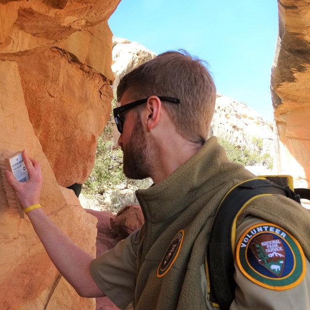 A person in a tan uniform and sunglasses brushing a sandstone wall.