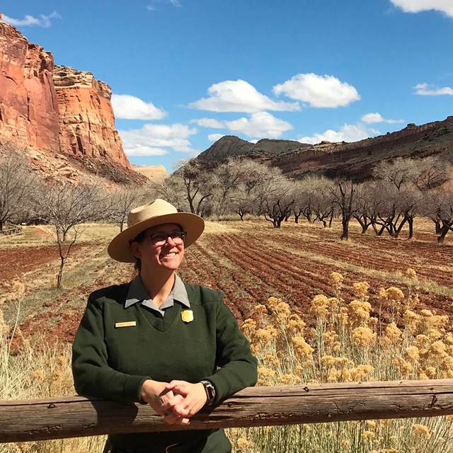 A ranger leaning on a wooden fence with rows of bare orchard trees and red cliffs in the background.