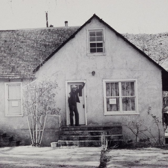 Black and white photo of a man standing on the porch of a house. 