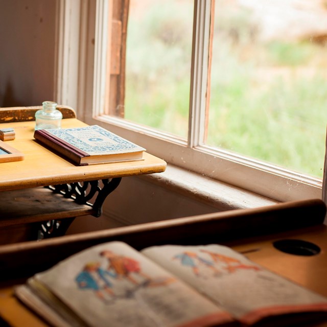 Photo of old-fashioned school books on old-fashioned desks beside windows.
