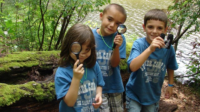 Three children smile as they hold magnifying glasses on a park trail