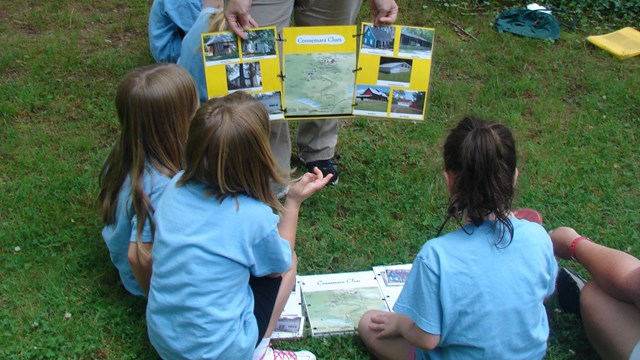 A group of children sit on the grass while an adult shows them a map.