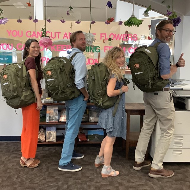 Four people hold green NPS backpacks while standing in a library. 