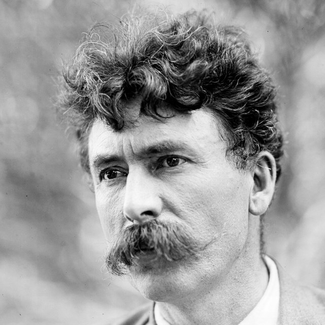 Closeup black & white historic portrait photo of a man with dark curly hair and a large mustache.