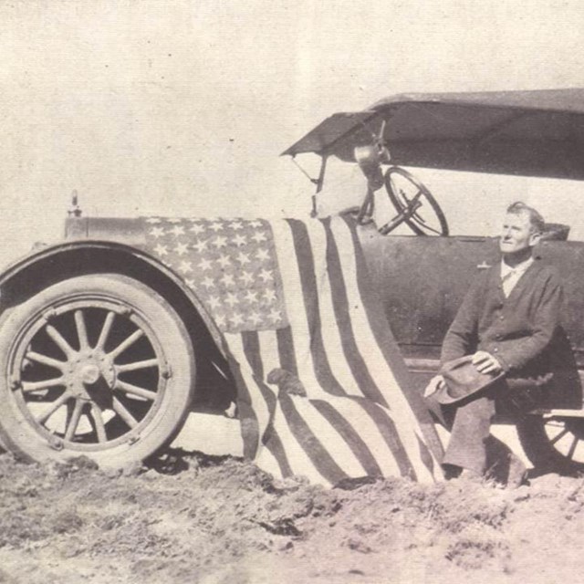A black and white photo of a man kneeling in front with an American flag draped over the hood.