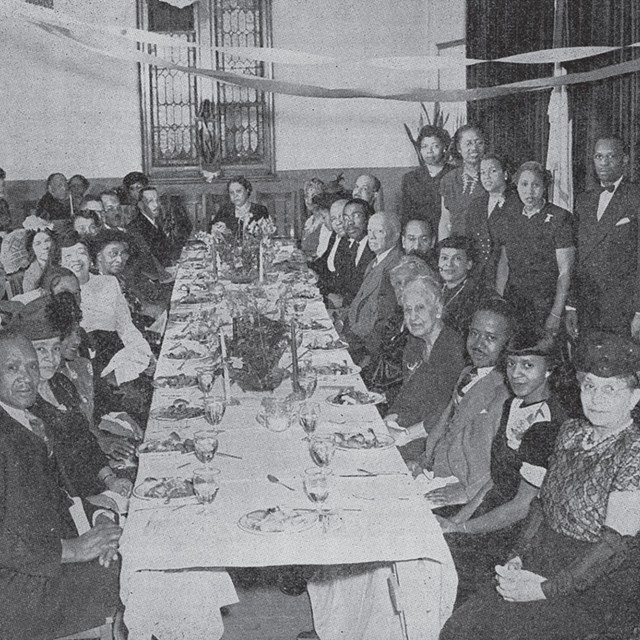 A large group of African Americans in formal dress sits around a long table