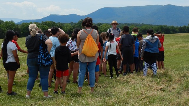 Park ranger leads a group of students across an open meadow. Large mountain rises up behind them.