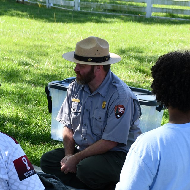 A park ranger sits on a lawn with 4th graders, showing them historic clothing.