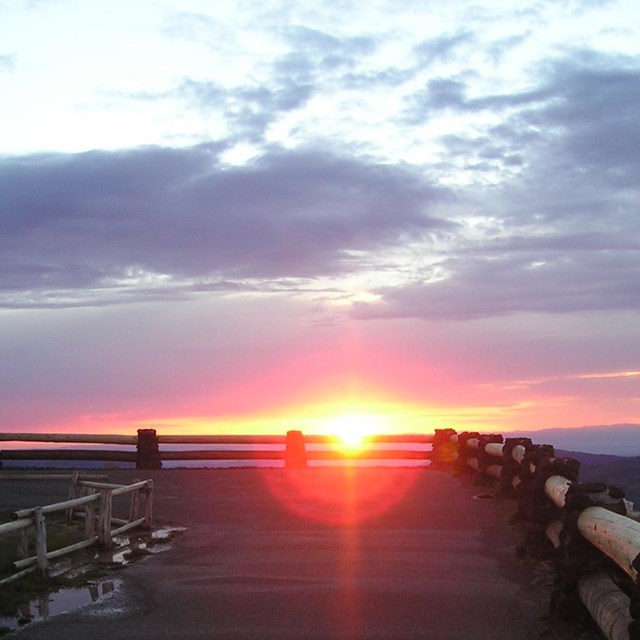 A wooden railing leads to an overlook, the sun sets in the distance.