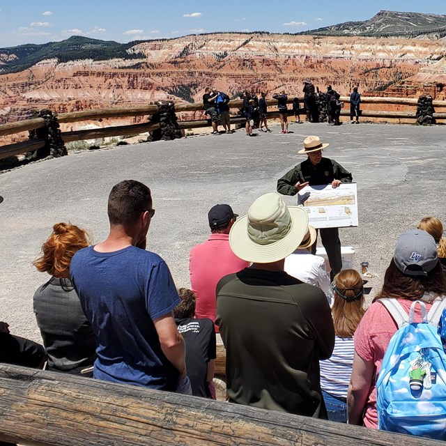 A large group is gathered around a ranger speaking with multicolored rock formations in the distance