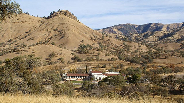 A white building with a red roof is situated among yellow grassy hills.