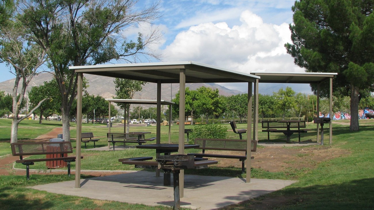 A group of three covered picnic tables with grills and trash cans