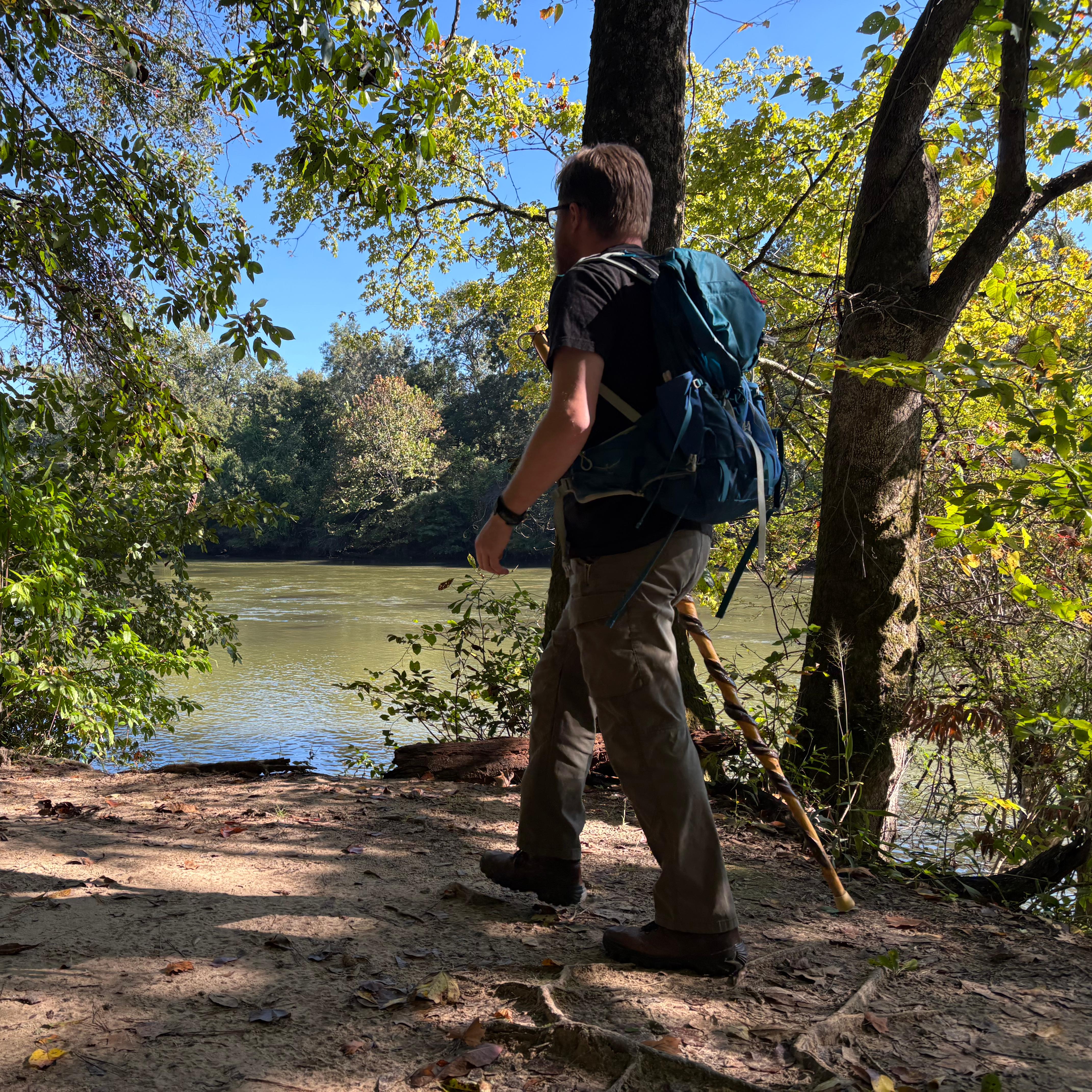 A man walks on a wooded dirt path with a blue green river flowing in the background. 