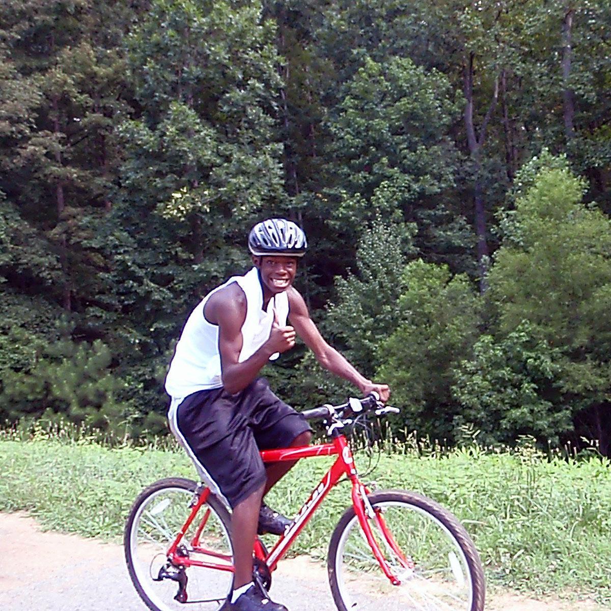 A young man rides a red bike on a paved trail while giving a 'thumbs up' sign.