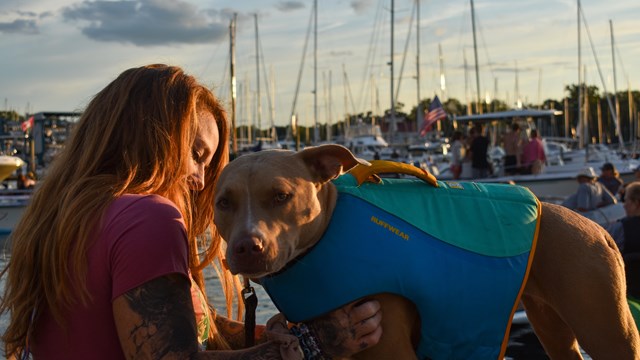 A woman sits on a dock facing her dog, who is facing the camera.