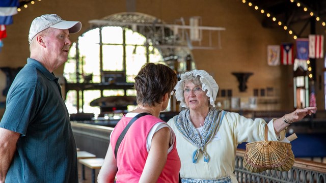 A woman in period clothing talks with a man in a woman in an indoor setting.