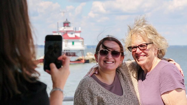 Two women pose for a picture in front of a lighthouse in the water in the distance behind them.