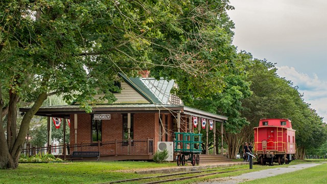 A red caboose sits on tracks next to a historic train station in a park setting.
