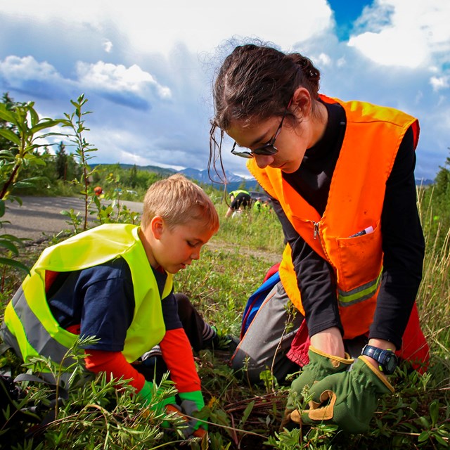 a young boy and girl in safety vests pull weeds