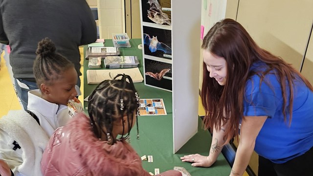 Intern in blue shirt showing two children educational materials about bats.