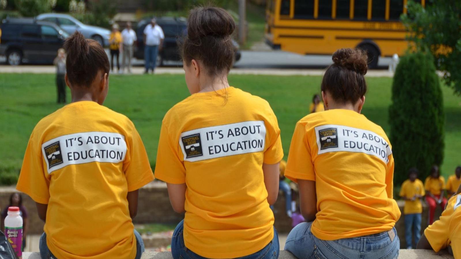 Three students in yellow shirts reading &ldquo;It&rsquo;s About Education&rdquo; sit together near a school bus.