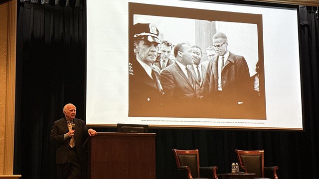 A man speaks in front of a podium with a photo of Dr. Martin Luther King in the background