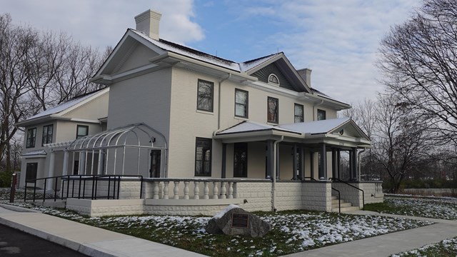 Two story historic brick home pained light gray surrounded by green grass with light snow on it all.