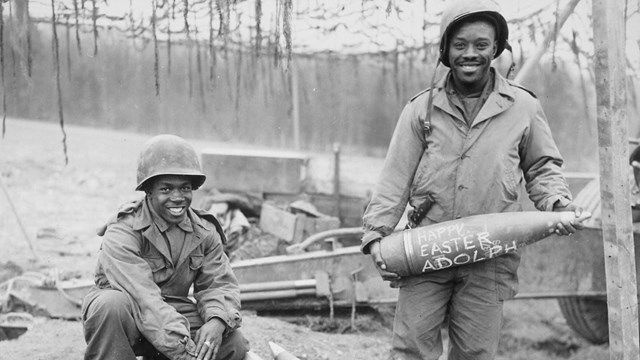 Two african Americans stand with artillery shells smiling at camera. They are wearing WW2 uniforms.