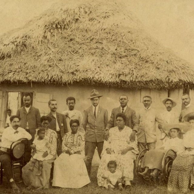 African civilians and African American soldiers in rows in front of a straw roof building