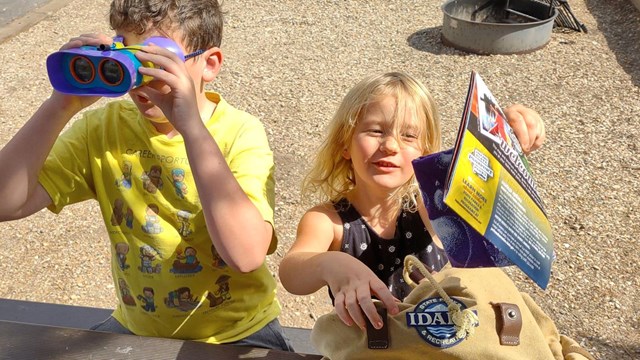 A boy in a yellow shirt with binoculars and a young girl pulling a booklet from a backpack.