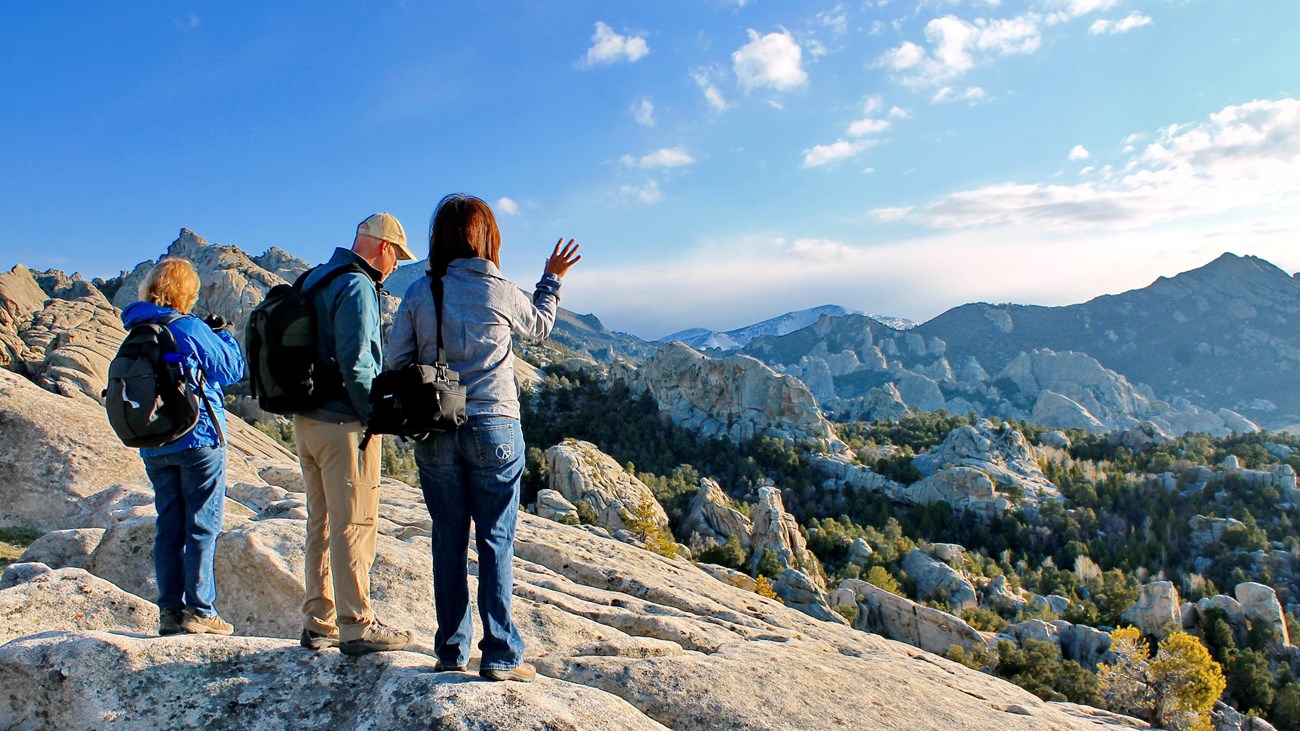 Three people standing on a granite slab look out over a grand scenic view of City of Rocks.