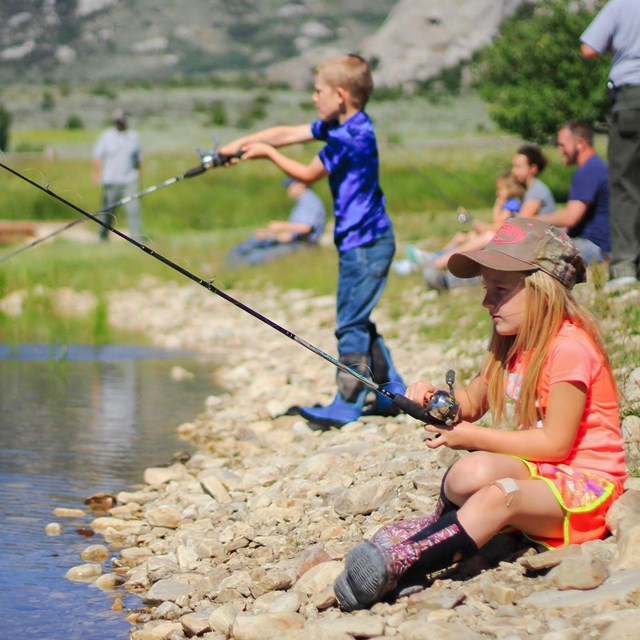 Kids and adults sit along a rocky pond shore fishing.