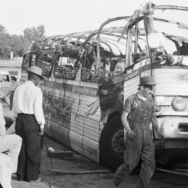 A tow truck tows a burned out bus that was bombed with African American and White men observing