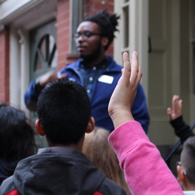 Kids raise their hands to ask questions of a presenter standing in front of a brick building