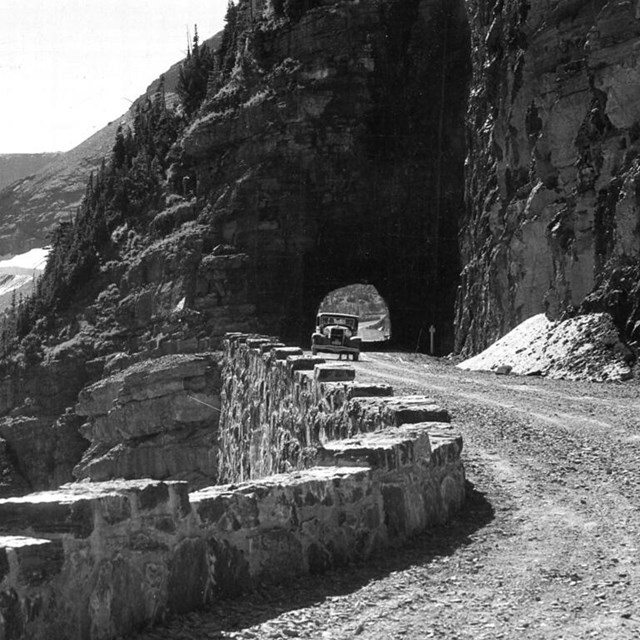 An old-fashioned car drives under a rock tunnel at Glacier National Park