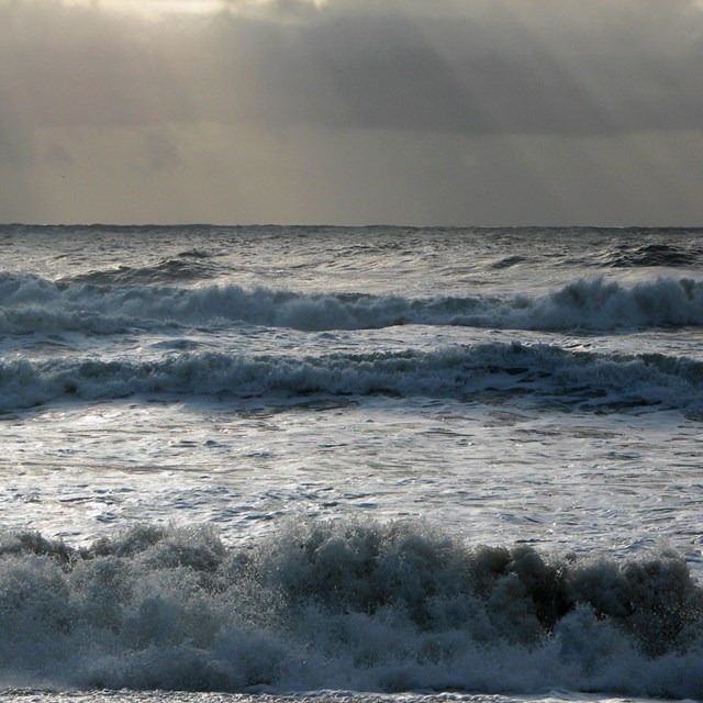 An ocean view looking directly out at white frothy waves with sunrays shining through brown clouds