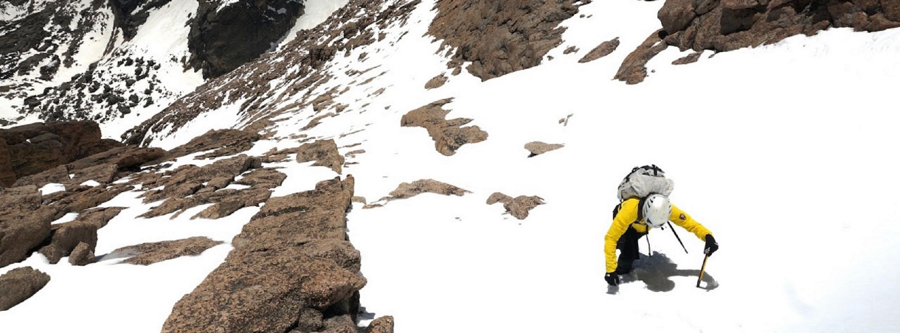 Park ranger ascending a steep snow field, mountains and trees far below.  